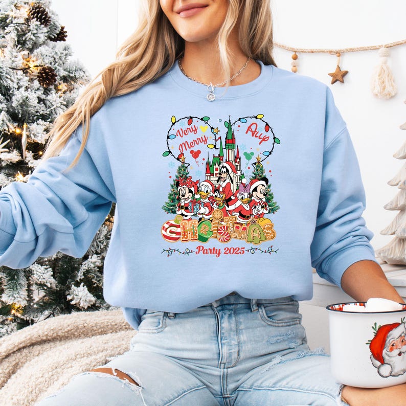 May include: Light blue sweatshirt featuring a colorful Christmas graphic with Disney characters. The design includes the text "Very Merry Christmas Party 2025" and a castle illustration. The sweatshirt has a crew neck and long sleeves.