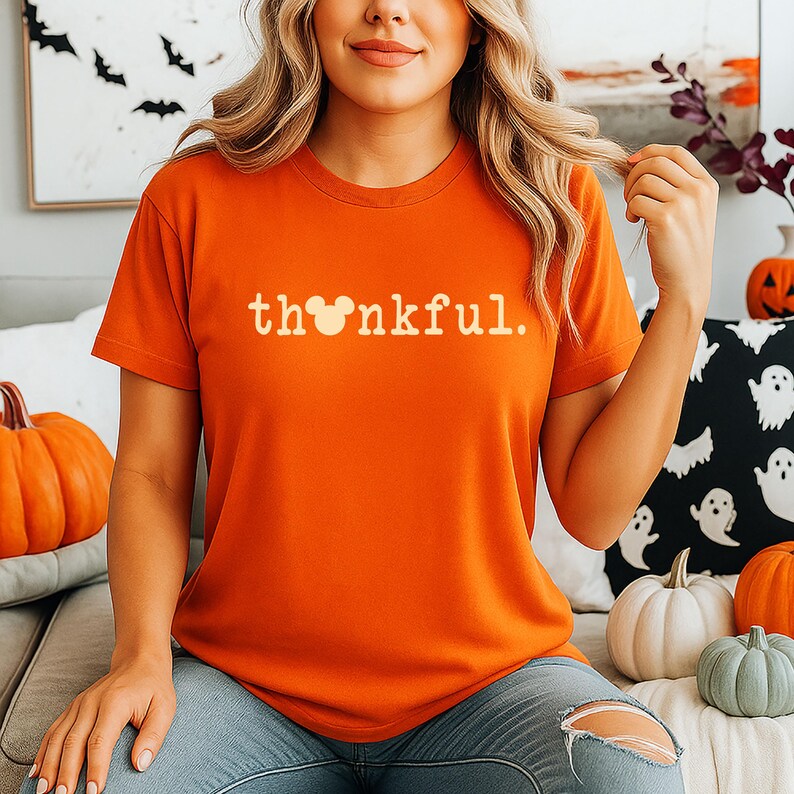 Puede incluir: Camiseta naranja con la palabra "thankful" en blanco, con una cabeza de Mickey Mouse reemplazando la "o". La camiseta es parte de una colecci&oacute;n con tem&aacute;tica de oto&ntilde;o o Halloween. Las calabazas y las almohadas decorativas est&aacute;n en el fondo.