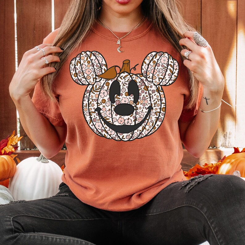 May include: A burnt orange t-shirt with a floral pumpkin Mickey Mouse design. The pumpkin has a smiling face and is surrounded by small white flowers.
