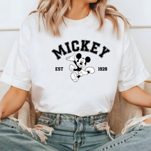 May include: White t-shirt with a black and white graphic of Mickey Mouse. The graphic includes the text "Mickey" in a black arch above Mickey Mouse, "EST" to the left of Mickey Mouse, and "1928" to the right of Mickey Mouse.