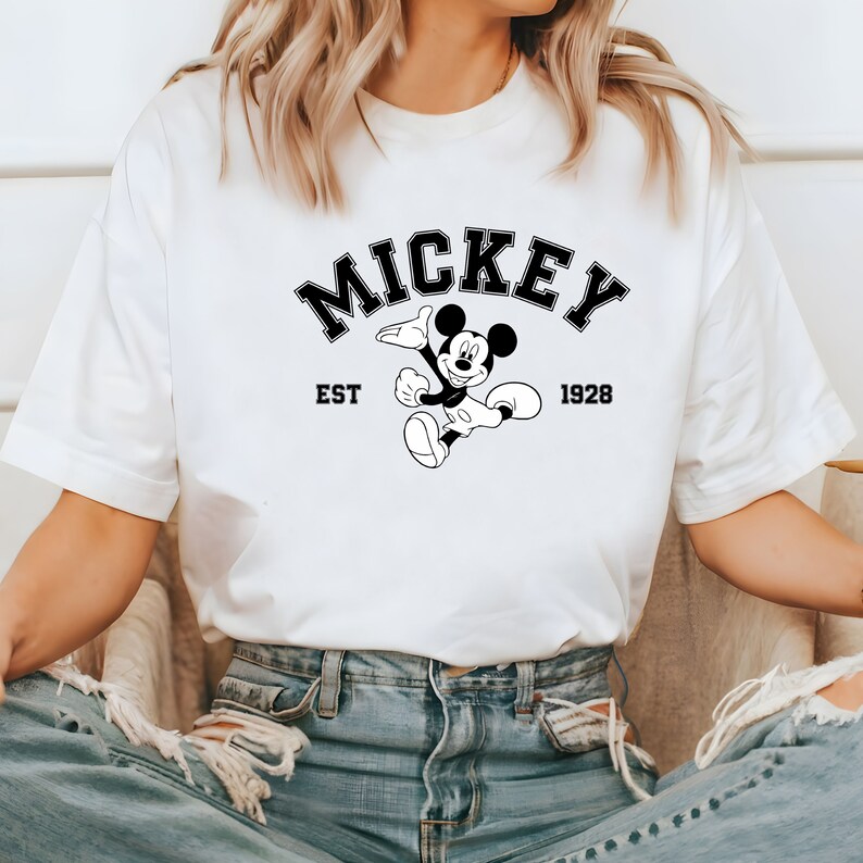 May include: White t-shirt with a black and white graphic of Mickey Mouse. The graphic includes the text "Mickey" in a black arch above Mickey Mouse, "EST" to the left of Mickey Mouse, and "1928" to the right of Mickey Mouse.