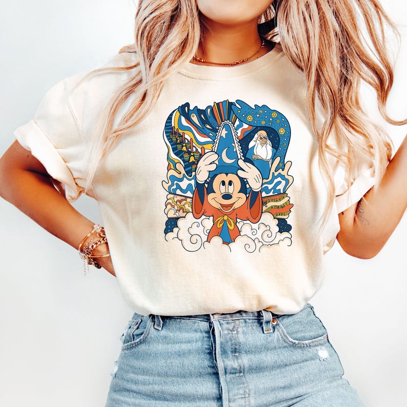 May include: A white t-shirt with a vintage-style illustration of Mickey Mouse as a sorcerer. The illustration features Mickey Mouse wearing a blue sorcerer's hat with a crescent moon and star, a red robe, and a yellow sash. The illustration also includes a background of clouds, stars, and a castle.