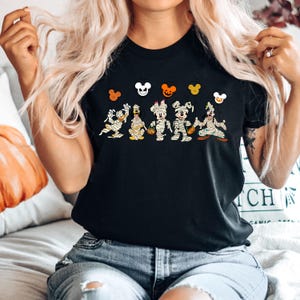 Peut inclure: T-shirt noir avec un motif de Mickey Mouse, Minnie Mouse, Donald Duck, Dingo et Pluto d&eacute;guis&eacute;s en momies pour Halloween. Les personnages tiennent des citrouilles et des bonbons. Il y a cinq t&ecirc;tes de Mickey Mouse au-dessus des personnages.