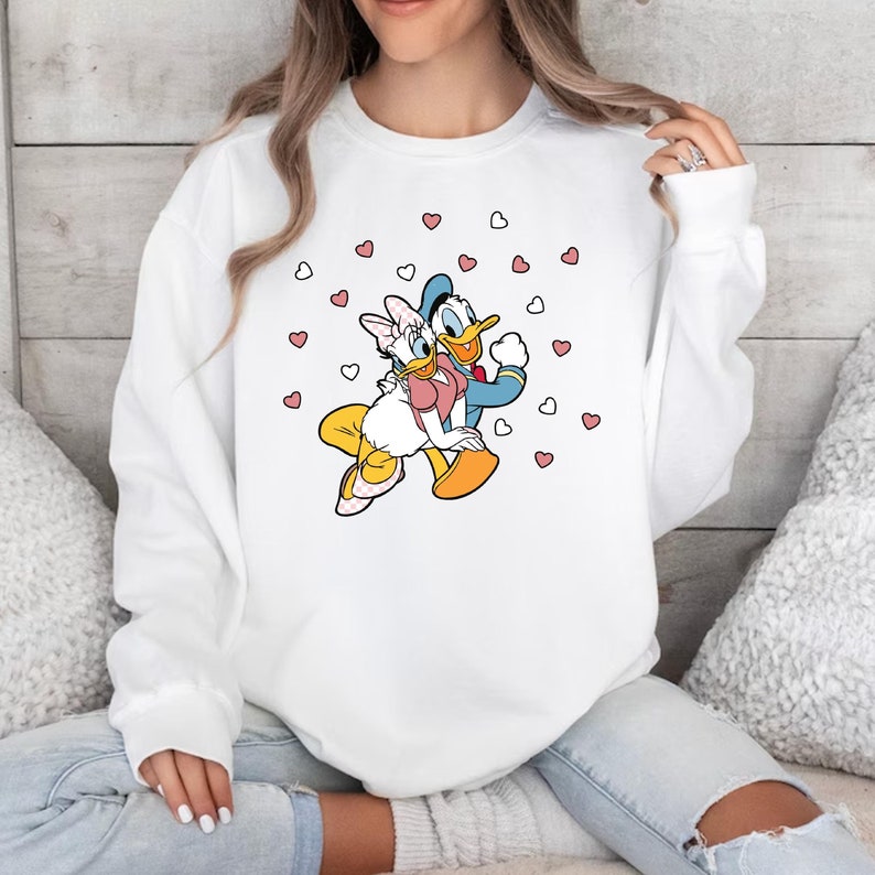 May include: White sweatshirt with a graphic of Donald Duck and Daisy Duck holding each other surrounded by pink hearts.
