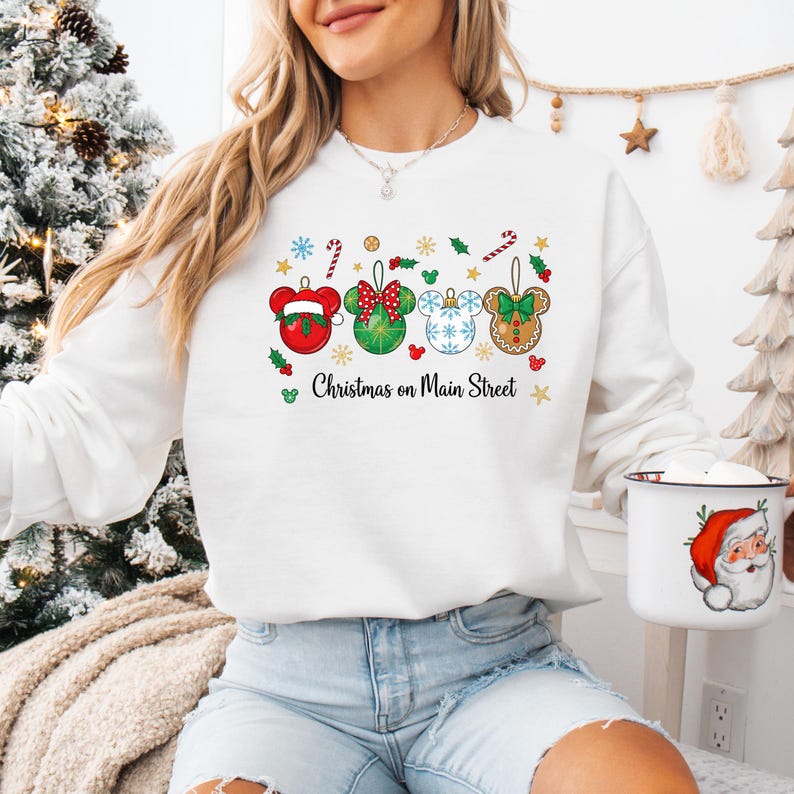 May include: White sweatshirt with a Christmas-themed design. The design features Mickey Mouse head-shaped Christmas ornaments in red, green, and blue, with the text "Christmas on Main Street". A mug with a Santa Claus graphic is held in the hand.