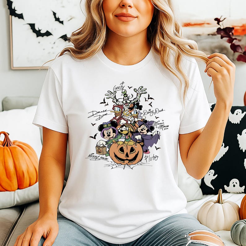 May include: White t-shirt featuring a Halloween-themed graphic of Disney characters, including Mickey Mouse, Donald Duck, and Goofy, surrounding a carved pumpkin. The design includes bats and character signatures.