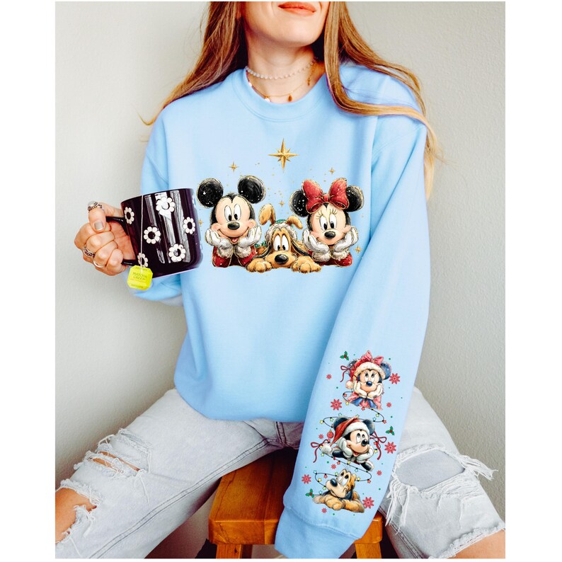 May include: Light blue sweatshirt featuring Mickey Mouse, Minnie Mouse, and Pluto. The design includes a star and festive accents. The sleeve displays additional Disney characters in holiday attire. A black mug with a floral design is also visible.