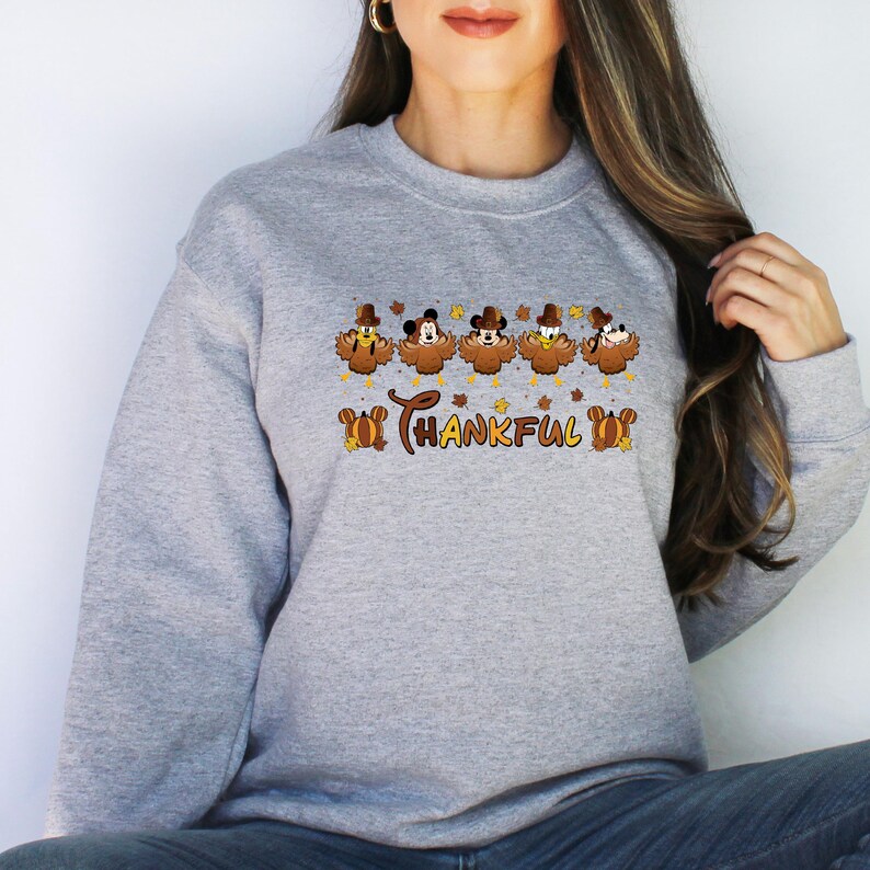 May include: A light gray sweatshirt featuring a Thanksgiving-themed design. The design includes cartoon turkey characters wearing hats, with the word "THANKFUL" in brown script. Autumn leaves are scattered around the text and characters.