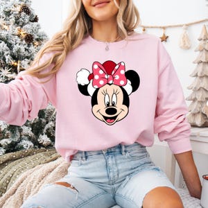 May include: Pink sweatshirt with a graphic of Minnie Mouse wearing a Santa hat and a red bow with white polka dots.