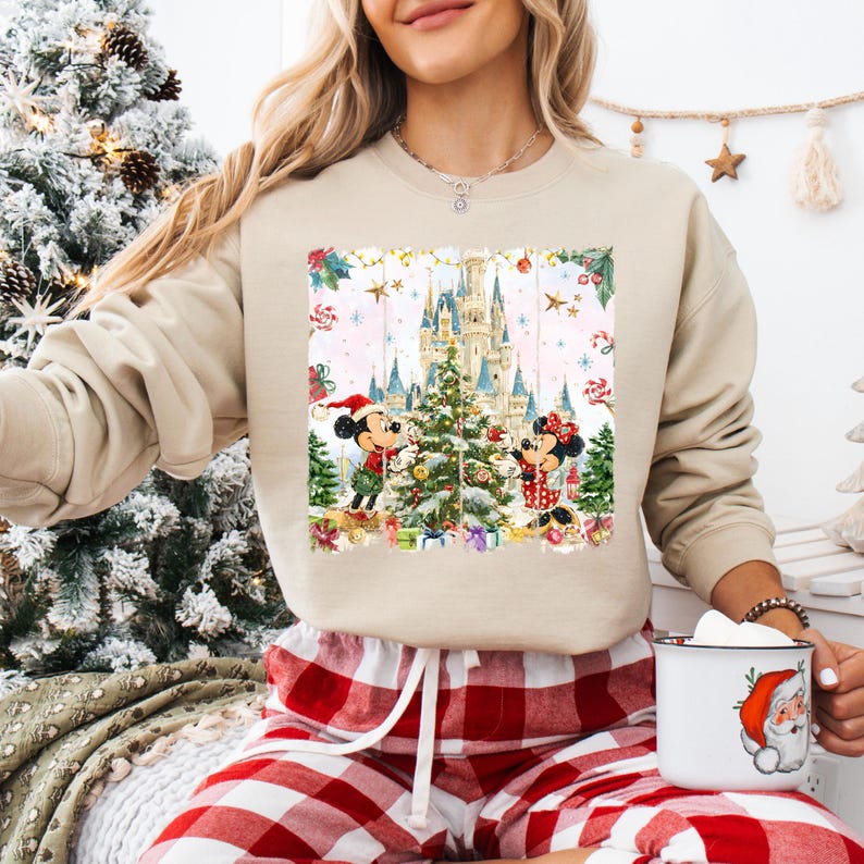 Może przedstawiać: Beżowa bluza z świąteczną grafiką Myszki Miki i Myszki Minnie przed zamkiem i choinką. Projekt zawiera prezenty i gwiazdki. Osoba ma na sobie czerwono-białą piżamę w kratę.