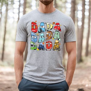 May include: A gray t-shirt with a graphic of the word "DADA" spelled out with cartoon cars from the movie Cars. Each letter is a different car, and the cars are all different colors.