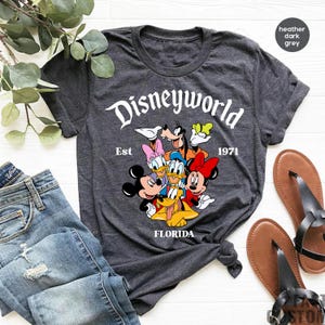 Pu&ograve; includere: T-shirt grigio scuro melange con una grafica bianca di Topolino, Minnie Mouse, Paperino, Pippo e Pluto. La grafica include il testo "Disneyworld Est 1971 Florida".