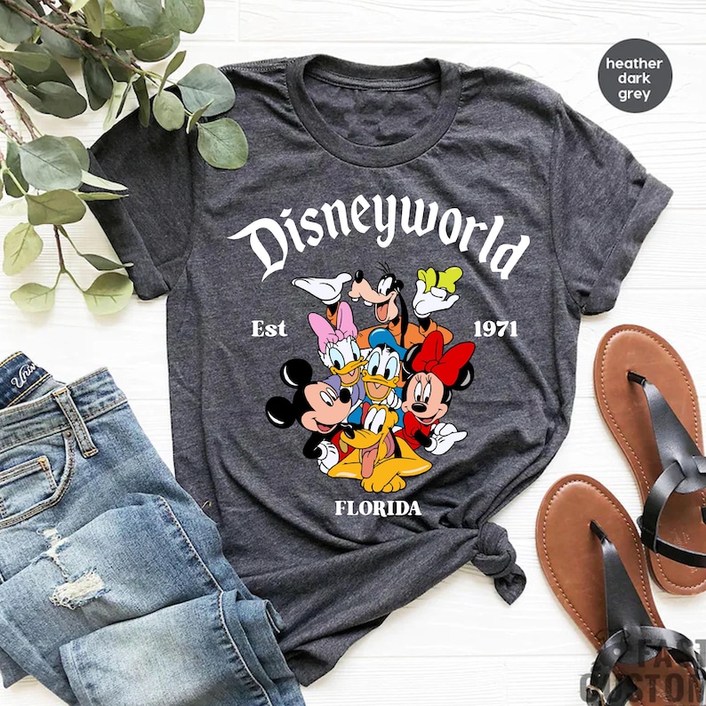 Pu&ograve; includere: T-shirt grigio scuro melange con una grafica bianca di Topolino, Minnie Mouse, Paperino, Pippo e Pluto. La grafica include il testo "Disneyworld Est 1971 Florida".