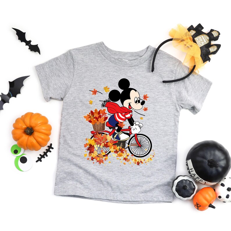 May include: A light gray t-shirt featuring Mickey Mouse riding a red bicycle through autumn leaves. Mickey wears a red scarf and carries a basket of leaves. Halloween decorations, including pumpkins and bats, surround the shirt.