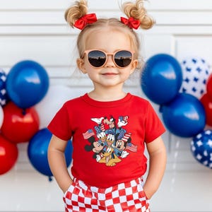 May include: A young child wearing a red t-shirt featuring Mickey Mouse, Donald Duck, Goofy, and other Disney characters with American flags. The child is also wearing sunglasses and red and white checkered shorts. Balloons in red, white, and blue are in the background.