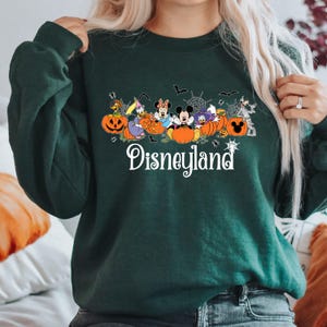 K&ouml;nnte beinhalten: Dunkelgr&uuml;nes Sweatshirt mit einem Halloween-Disneyland-Design. Die Grafik zeigt Disney-Figuren, K&uuml;rbisse, Flederm&auml;use und Spinnweben. Das Wort "Disneyland" steht in wei&szlig;er Schrift am unteren Rand.