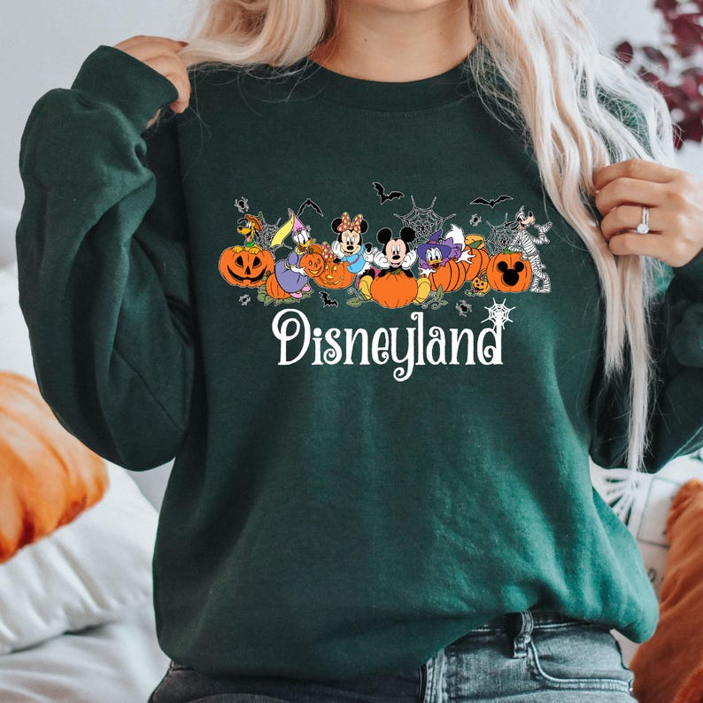 K&ouml;nnte beinhalten: Dunkelgr&uuml;nes Sweatshirt mit einem Halloween-Disneyland-Design. Die Grafik zeigt Disney-Figuren, K&uuml;rbisse, Flederm&auml;use und Spinnweben. Das Wort "Disneyland" steht in wei&szlig;er Schrift am unteren Rand.