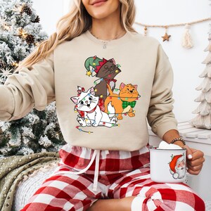 May include: Beige sweatshirt featuring a graphic of three cartoon cats adorned with Christmas lights and festive accessories. The cats are white, brown, and orange. The sweatshirt is a neutral color, suitable for the holiday season.