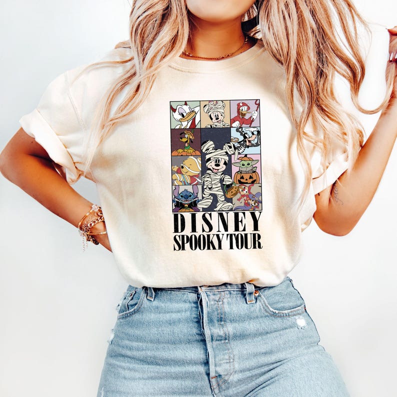 Pu&ograve; includere: T-shirt color crema con una grafica colorata di personaggi Disney in costume di Halloween. La grafica &egrave; divisa in quadrati e include personaggi come Topolino, Paperino e Stitch. Il testo "DISNEY SPOOKY TOUR" &egrave; stampato sotto.