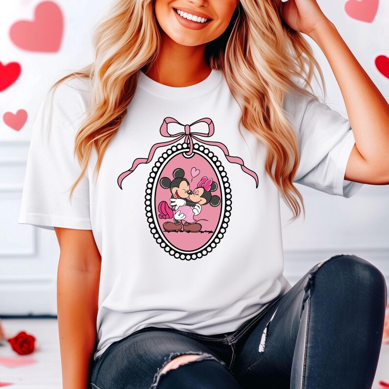 K&ouml;nnte beinhalten: Wei&szlig;es T-Shirt mit einer rosa Schleife und einem rosa ovalen Rahmen, in dem Mickey Mouse und Minnie Mouse sich k&uuml;ssen. Der Rahmen ist mit schwarzen und wei&szlig;en Punkten verziert.
