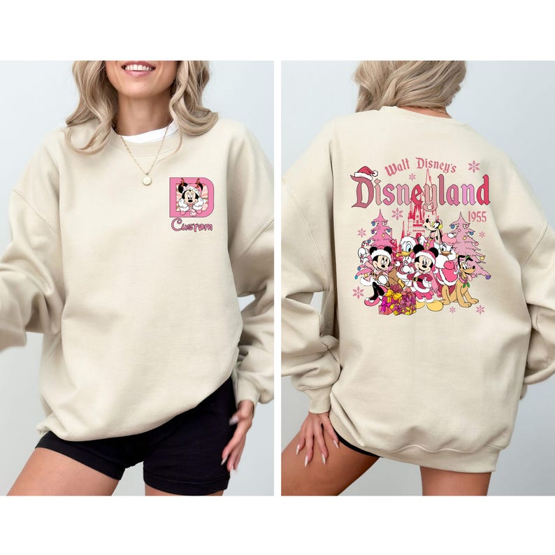 May include: Beige sweatshirt featuring a pink Minnie Mouse design inside a pink letter "D" on the front. The back displays a Disneyland graphic with characters, trees, and text: "Walt Disney's Disneyland 1955".