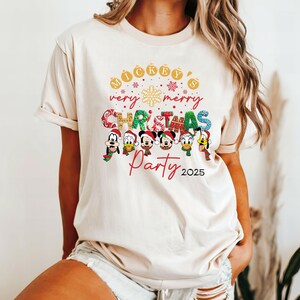 K&ouml;nnte beinhalten: Ein beiges T-Shirt mit einem festlichen Weihnachtsmotiv mit Mickey Mouse und seinen Freunden. Der Text lautet "Mickey's very merry Christmas Party 2025".