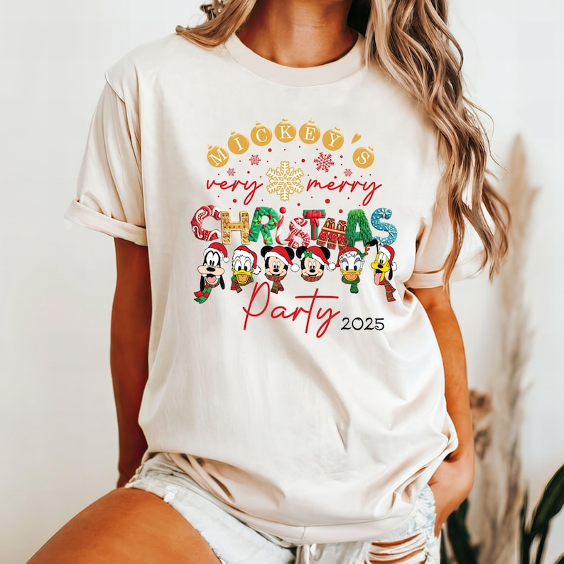 K&ouml;nnte beinhalten: Ein beiges T-Shirt mit einem festlichen Weihnachtsmotiv mit Mickey Mouse und seinen Freunden. Der Text lautet "Mickey's very merry Christmas Party 2025".