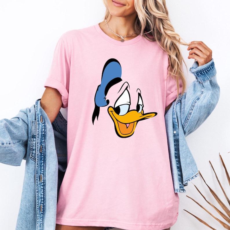 May include: A light pink t-shirt featuring a cartoon image of Donald Duck's face. The character's face is in the center of the shirt, with a blue hat, yellow beak, and black outlines. The shirt has short sleeves and a crew neck.