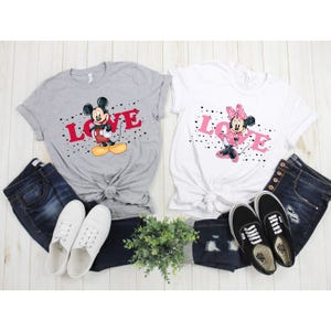 Op de afbeelding: Twee grijze en witte T-shirts met een Mickey Mouse en Minnie Mouse afbeelding. De shirts tonen het woord "LOVE" in rode letters en zwarte hartjes. De shirts liggen op een witte ondergrond met jeans en schoenen.