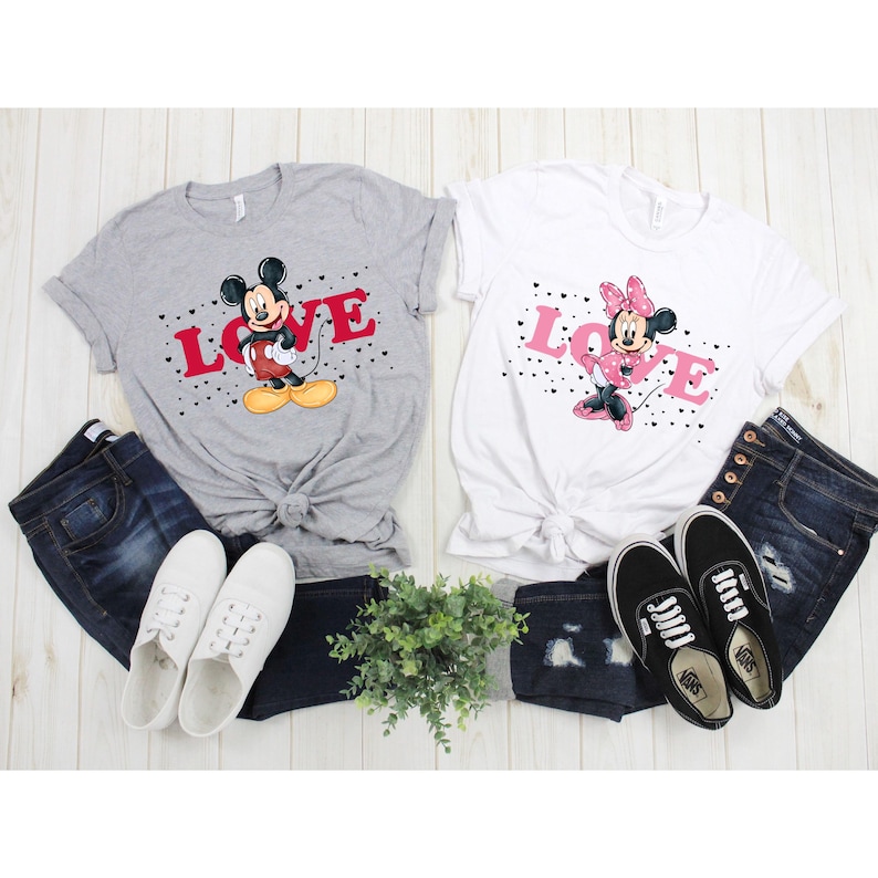 Op de afbeelding: Twee grijze en witte T-shirts met een Mickey Mouse en Minnie Mouse afbeelding. De shirts tonen het woord "LOVE" in rode letters en zwarte hartjes. De shirts liggen op een witte ondergrond met jeans en schoenen.