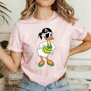 May include: A light pink t-shirt featuring a cartoon duck dressed as a pirate. The duck wears a black eye patch, a pirate hat with a skull and crossbones, and a green and white striped top. The shirt has rolled-up sleeves.