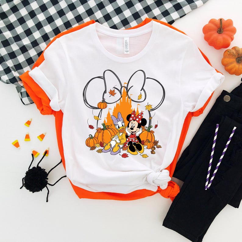 May include: White t-shirt with orange trim featuring a Minnie Mouse and Daisy Duck design surrounded by pumpkins and autumn leaves. The shirt is paired with black pants, candy corn, and decorative pumpkins.