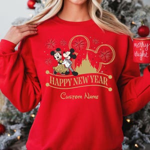 May include: Red sweatshirt with a gold Mickey Mouse and Minnie Mouse design. The design includes fireworks, a castle, and the text "Happy New Year". The text "Custom Name" is also included on the sweatshirt.