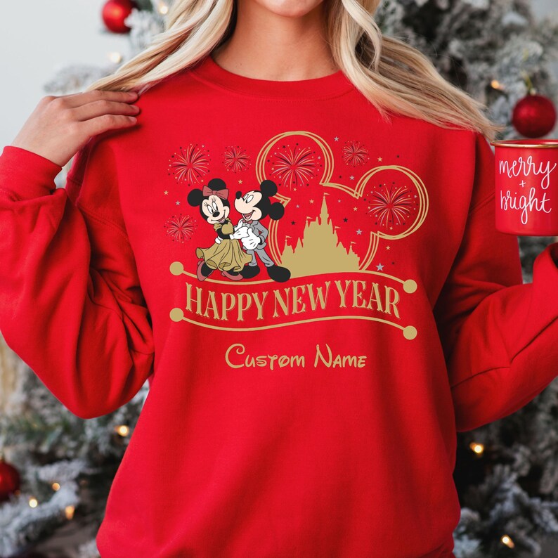 May include: Red sweatshirt with a gold Mickey Mouse and Minnie Mouse design. The design includes fireworks, a castle, and the text "Happy New Year". The text "Custom Name" is also included on the sweatshirt.
