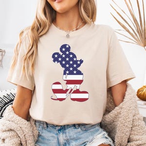 May include: Beige t-shirt with a Mickey Mouse silhouette filled with a patriotic American flag design. The design includes white stars on a blue background for the head and red and white stripes for the body. The shirt has short sleeves.