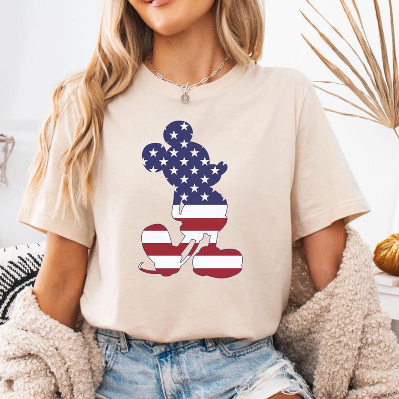 May include: Beige t-shirt with a Mickey Mouse silhouette filled with a patriotic American flag design. The design includes white stars on a blue background for the head and red and white stripes for the body. The shirt has short sleeves.