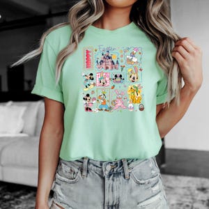 May include: A light green t-shirt featuring a colorful graphic with Disney characters and the words "Happy Easter". The graphic includes Mickey and Minnie Mouse, Donald Duck, Goofy, and other characters.