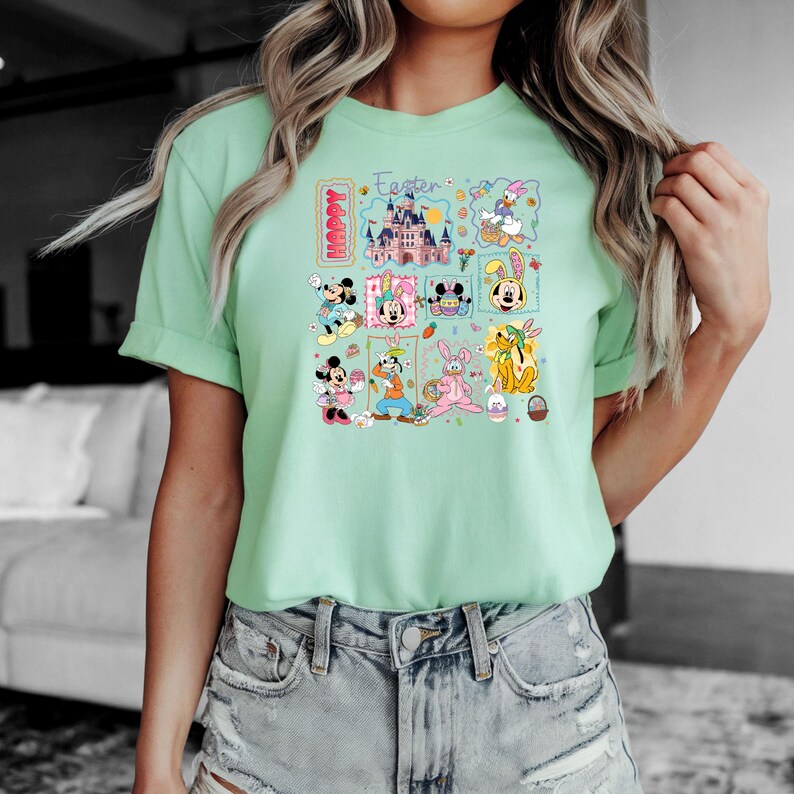 May include: A light green t-shirt featuring a colorful graphic with Disney characters and the words "Happy Easter". The graphic includes Mickey and Minnie Mouse, Donald Duck, Goofy, and other characters.