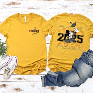 以下が含まれることがあります： 後ろにミッキーマウスのグラフィックが描かれた黄色のTシャツ2枚。1枚のシャツの後ろには「Senior 2025」と書いてあり、もう1枚のシャツの前には「Senior 2025 Johan」と書いてあります。