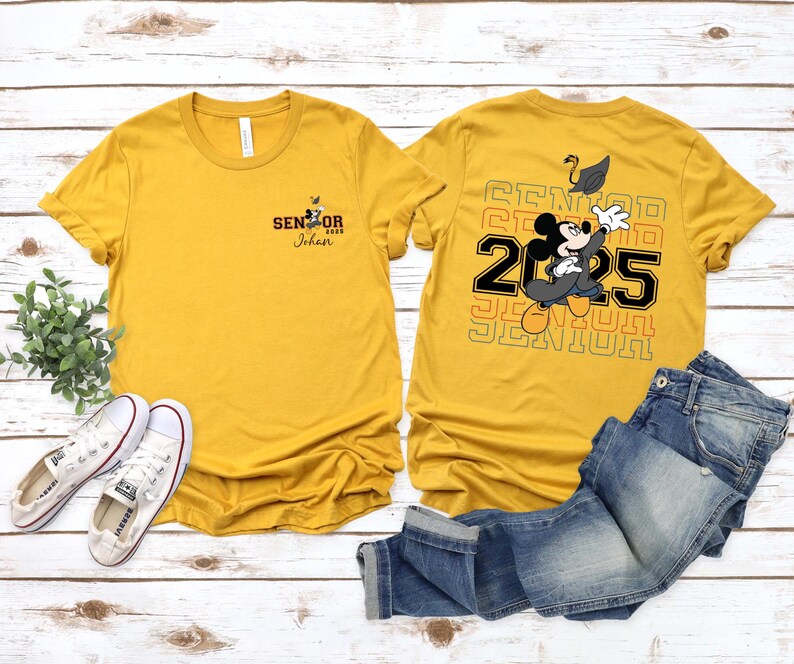 以下が含まれることがあります： 後ろにミッキーマウスのグラフィックが描かれた黄色のTシャツ2枚。1枚のシャツの後ろには「Senior 2025」と書いてあり、もう1枚のシャツの前には「Senior 2025 Johan」と書いてあります。