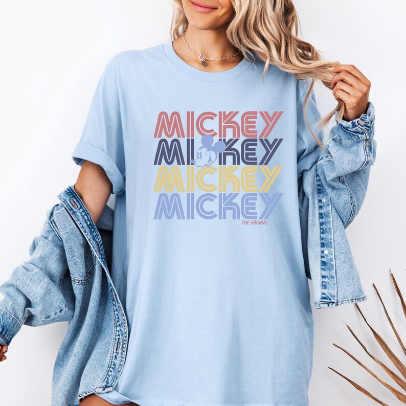 May include: Light blue t-shirt with the word "MICKEY" stacked four times in different colors. The word "MICKEY" is in red, purple, yellow, and blue. A small Mickey Mouse silhouette is in the second "MICKEY". The text "THE ORIGINAL" is below the last "MICKEY".