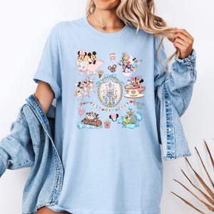 May include: Light blue t-shirt featuring a colorful graphic with various Disney characters and the text "Best Day Ever." The design includes Mickey and Minnie Mouse, a castle, and other iconic elements from Disney movies.