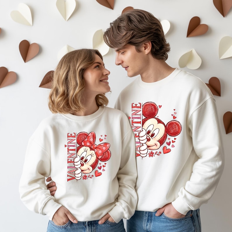 May include: Matching white sweatshirts with Mickey and Minnie Mouse designs for Valentine's Day. The sweatshirts have red accents, including hearts and the word "VALENTINE" printed vertically. The couple is standing in front of a wall decorated with paper hearts.