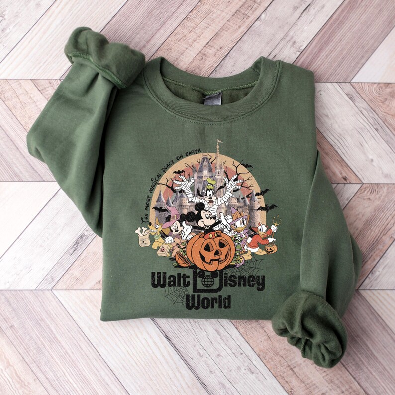 Puede incluir: Sudadera verde con un dise&ntilde;o de Halloween con personajes de Disney como Mickey Mouse y Donald Duck. El dise&ntilde;o incluye una calabaza, un castillo y el texto "Walt Disney World". Tambi&eacute;n incluye la frase "The most magical place on Earth".