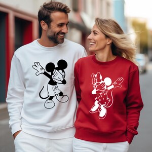 May include: A couple wearing matching sweatshirts with a white and black Mickey Mouse and Minnie Mouse graphic. The man is wearing a white sweatshirt and the woman is wearing a red sweatshirt.