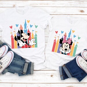 May include: Two white t-shirts with a colorful castle silhouette and Mickey Mouse and Minnie Mouse characters. The shirts are laid out on a white wooden surface with blue jeans and white sneakers.
