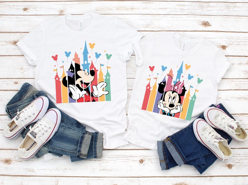 May include: Two white t-shirts with a colorful castle silhouette and Mickey Mouse and Minnie Mouse characters. The shirts are laid out on a white wooden surface with blue jeans and white sneakers.