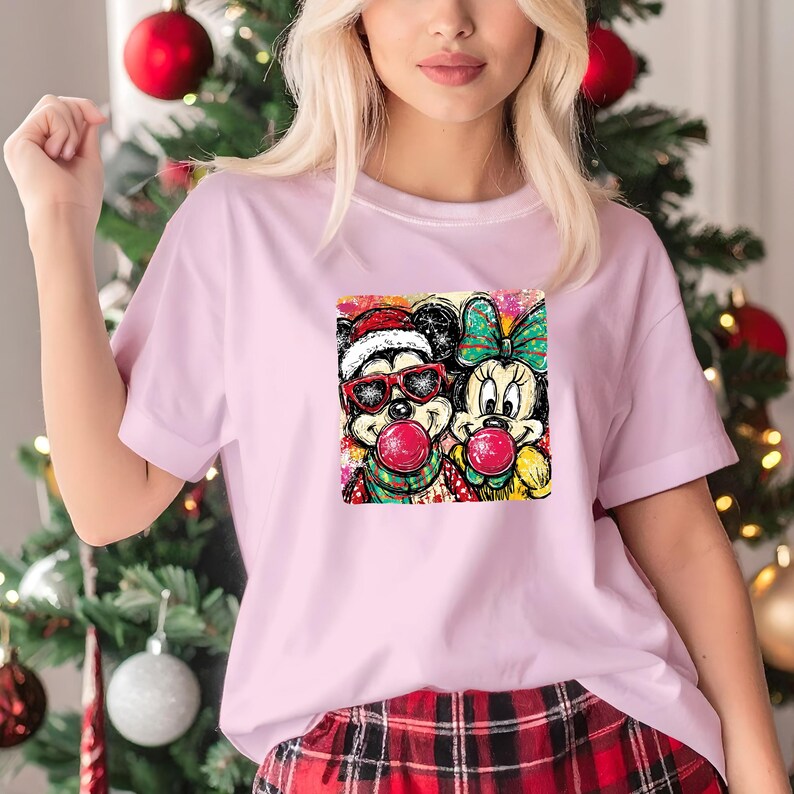 May include: A light pink t-shirt featuring a colorful graphic of Mickey and Minnie Mouse blowing bubble gum. Mickey wears a Santa hat and sunglasses, while Minnie has a bow. The shirt is paired with plaid pants.