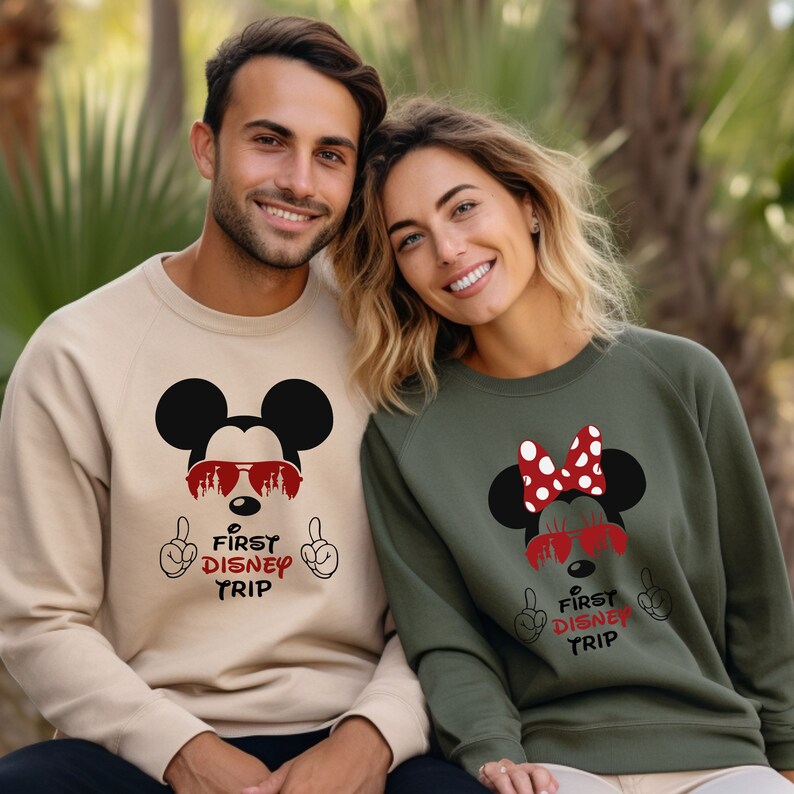 Op de afbeelding: Een stel draagt ​​passende sweatshirts met een Mickey Mouse en Minnie Mouse ontwerp. De sweatshirts zijn beige en groen en bevatten de tekst "First Disney Trip" met Mickey Mouse en Minnie Mouse die een zonnebril dragen.