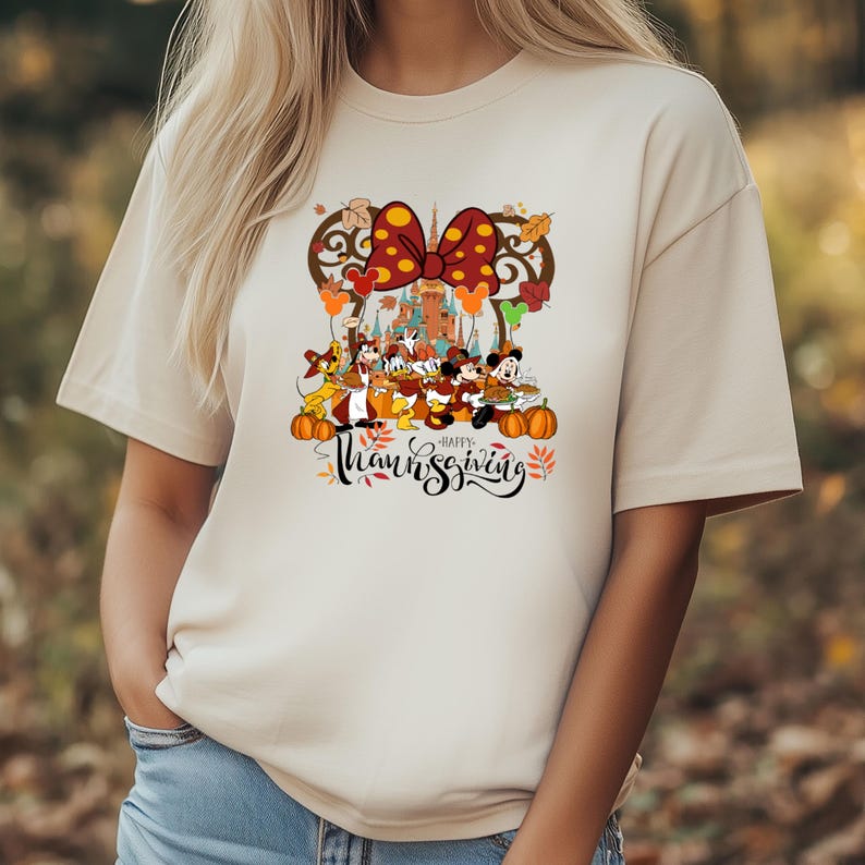 May include: Beige t-shirt featuring a colorful graphic of Disney characters celebrating Thanksgiving. The design includes Mickey and Minnie Mouse, a castle, pumpkins, and the words "Happy Thanksgiving" in a script font.